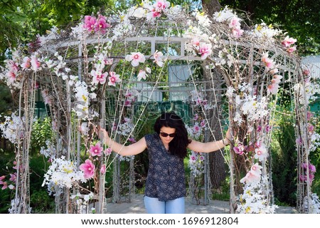Brunette girl in a white arbor made of artificial pink flowers. Decorative arbor girl in black glasses poses for the camera.