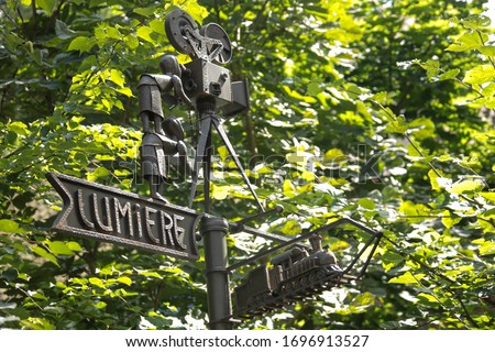Cast-iron direction indicator on a background of green wood. The arrow says "Lumiere" and the old train. From above, two men are looking at the old movie camera.