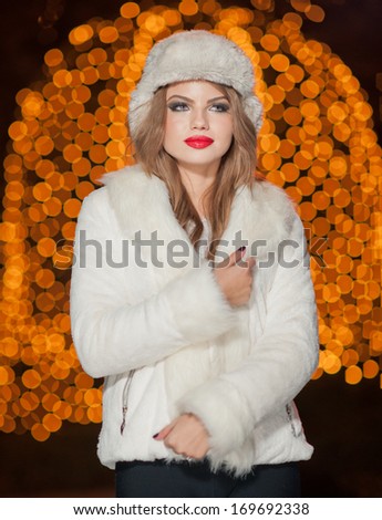 Fashionable lady wearing white fur cap and coat outdoor with bright Xmas lights in background. Portrait of young beautiful woman in winter style. Bright picture of beautiful blonde woman with make up