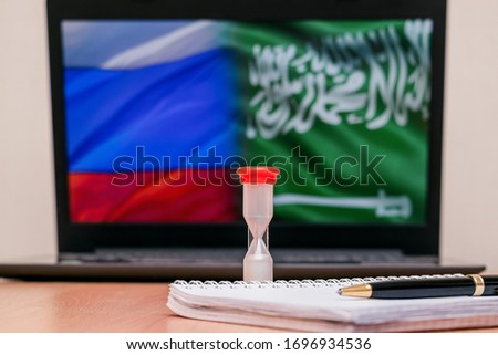 Hourglass, pen and notepad on the table.Blurry laptop with the flags of Russia and Saudi Arabia on the screen saver in the background.Concept of the OPEC meeting on April 9, 2020.Selective focus.