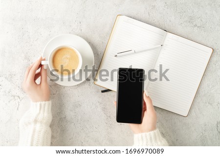 Beautiful women's hands holding up-to-date smartphone and writing in notepad with cup of coffee on concrete background. Women's business concept. Coffee break top view. Mockup for your design.