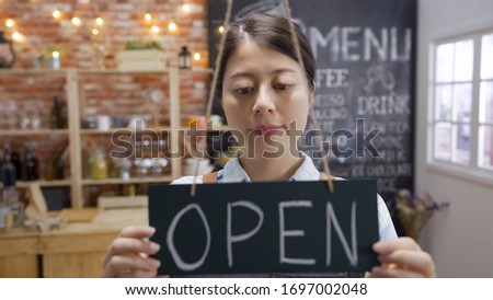 happy waitress standing at restaurant entrance flipping door plate. Portrait of mature business woman welcome customers in coffee shop in morning. aisan owner showing open sign in small business cafe