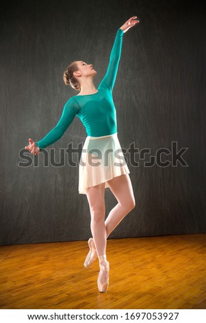 Young woman ballet dancer in a green leotard with tan skirt, dancing on pointe in a studio shot, against a gray background.