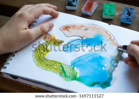 Child's drawing of watercolor paints in a sketch notebook, children's creativity concept. Child's hands next to the picture. Monotype. Drawing of an unusual fairytale animal.