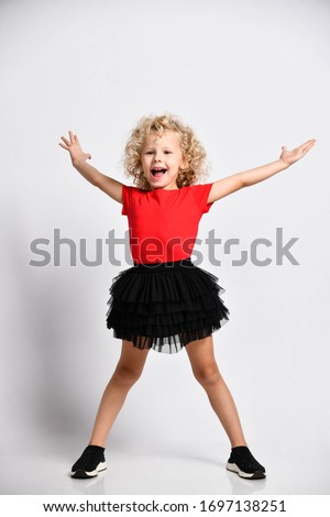 Cheerful happy screaming curly hair blonde kid girl in red t-shirt and black skirt poses with her legs wide apart and holding hands up