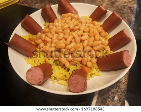 Deep round bowl of rice and baked beans with sliced juicy frankfurters around it 