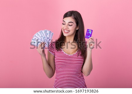 Photo of caucasian brunette woman 20s smiling while holding credit card and fan of dollar cash isolated over pink background
