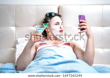 Funny man with a cosmetic mask on his face and curlers does spa treatments in a room holding cocktail in his hand spends a stream for social networks. Parody on cosmetics beauty and fashion bloggers Royalty-Free Stock Photo #1697265244