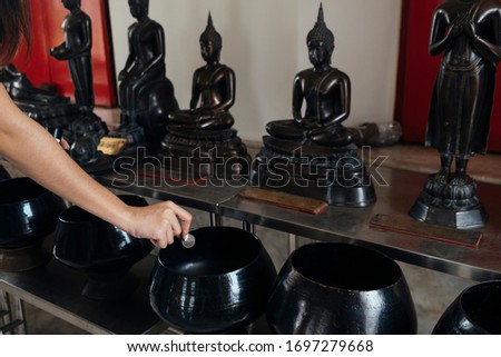 Cropped image of woman's hand drop the coin in the monk's alms-bowl on the table in front of Buddha statue.