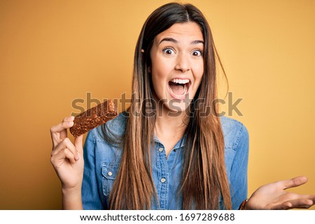 Young beautiful girl holding healthy protein bar standing over isolated yellow background very happy and excited, winner expression celebrating victory screaming with big smile and raised hands