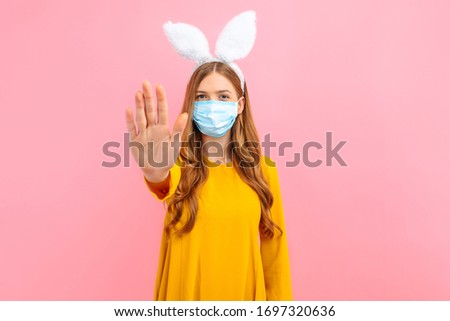 A young girl in the ears of an Easter Bunny, wears a medical mask on her face against viral diseases and shows a stop gesture, on an isolated pink background