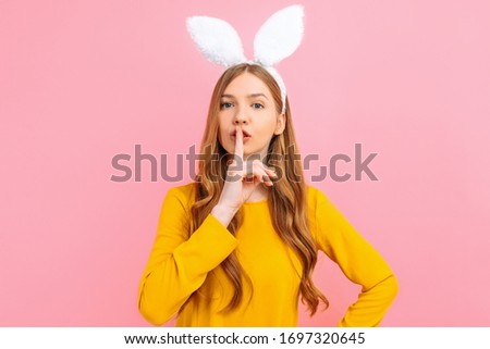 A young beautiful girl in the ears of an Easter Bunny, holds her front finger over her lips, makes a gesture of silence, speaks in a lower voice, on an isolated pink background