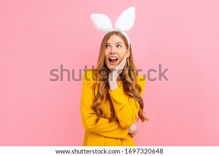 Happy surprised shocked young woman in Easter Bunny ears, looking at a copy of the space isolated on a pink background.
