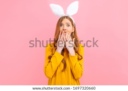 A shocked, terrified young woman in Easter Bunny ears isolated on a pink background.