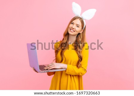 happy Easter. Happy young woman in the ears of an Easter Bunny holding a basket of colorful Easter eggs using a laptop on an isolated pink background