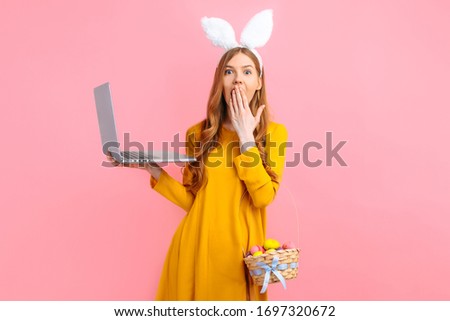 happy Easter. A shocked young woman in the ears of an Easter Bunny holding a basket of multicolored Easter eggs using a laptop on an isolated pink background