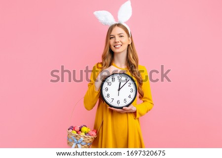 happy Easter. A beautiful girl in Easter Bunny ears holds a basket of colorful Easter eggs while holding a wall clock, on an isolated pink background