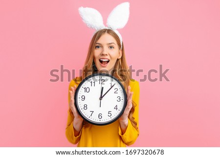 happy Easter. Beautiful girl in Easter Bunny ears holding a wall clock in her hands, on an isolated pink background