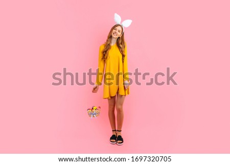 Happy Easter. A beautiful happy young woman wears rabbit ears on Easter day and holds a basket of Easter eggs, on an isolated pink background. Beautiful girl is getting ready for Easter