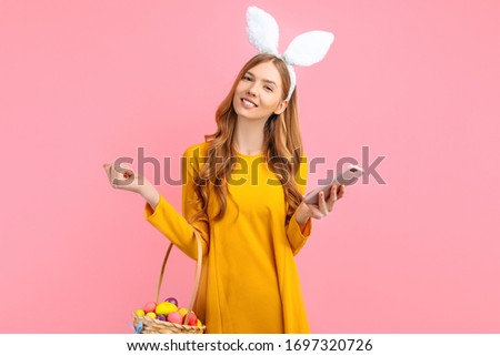 happy Easter. A happy young woman in the ears of an Easter Bunny, holding a basket of colorful Easter eggs, with a mobile phone in her hands on an isolated pink background