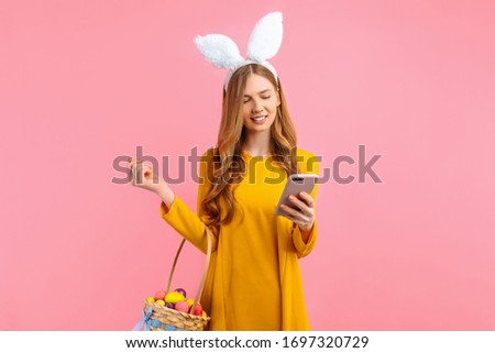 happy Easter. A happy young woman in the ears of an Easter Bunny, holding a basket of colorful Easter eggs, with a mobile phone in her hands on an isolated pink background