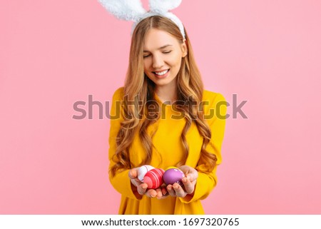 The concept of a happy Easter. A happy young girl with rabbit ears holds colorful Easter eggs, on a pink background.