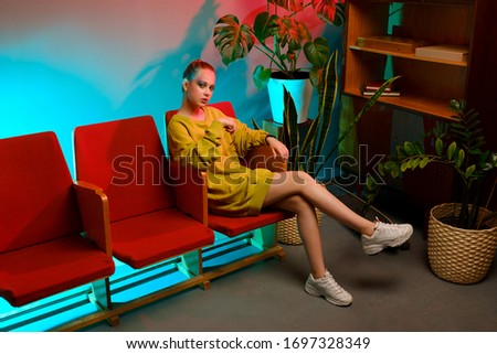 A young attractive girl with bright makeup and in colored clothes sits on a red armchair against a fashionable neon background. The interior of a modern teenager - a lot of flowers and longboards