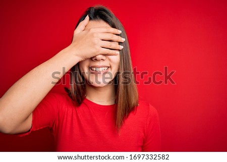 Young beautiful brunette girl wearing casual t-shirt over isolated red background smiling and laughing with hand on face covering eyes for surprise. Blind concept.