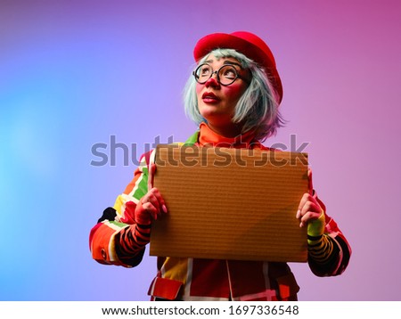 A clown girl with makeup, blue hair, in a red hat, a colored checkered jacket and glasses holds in her hands a blank cardboard for inscription. April Fools Day concept.