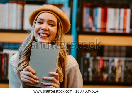 Portrait of young blonde woman posing in front of camera at book store while holding book with grey covers. Young girl is book lover. Royalty-Free Stock Photo #1697339614