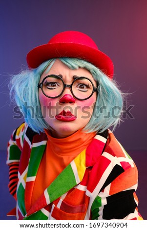 Close-up portrait of a clown girl with make-up, blue hair, a red hat, a colored checkered jacket and glasses. A clown shows different human emotions. April Fools Day concept. 