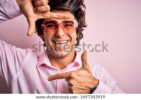 Young handsome man wearing glasses with heart form over isolated pink background smiling making frame with hands and fingers with happy face. Creativity and photography concept.