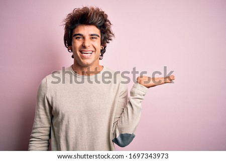 Young handsome man wearing casual t-shirt standing over isolated pink background smiling cheerful presenting and pointing with palm of hand looking at the camera.
