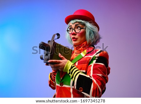 Close-up portrait of a clown girl with make-up, blue hair, a red hat, a colored checkered jacket and glasses. The clown is holding an old movie projector. April Fools Day concept.