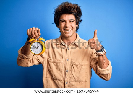 Young handsome man holding alarm clock standing over isolated blue background happy with big smile doing ok sign, thumb up with fingers, excellent sign