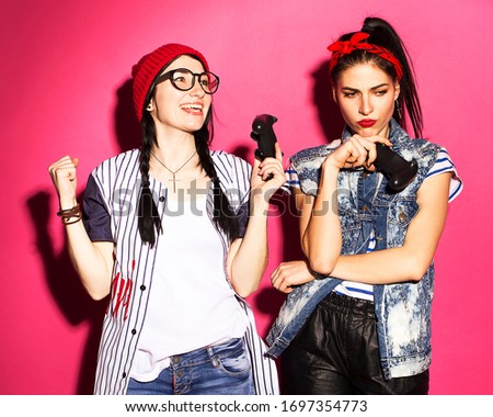 Two caucasian brunette hipster woman in casual stylish outfit having fun playing on console game using joysticks. They fight each other on a bright pink background
