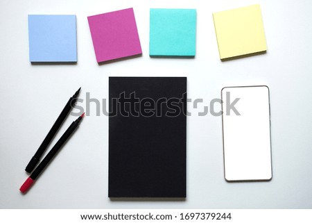 Top view of black book, pens, smart phone with white screen and label sheets or post note sticker set on white background