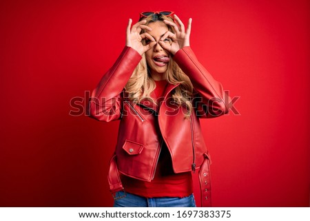 Young beautiful blonde woman wearing casual jacket standing over isolated red background doing ok gesture like binoculars sticking tongue out, eyes looking through fingers. Crazy expression.