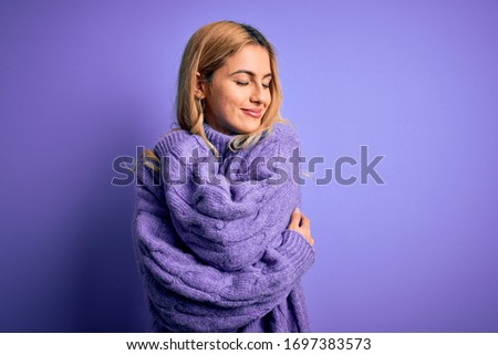 Young beautiful blonde woman wearing casual turtleneck sweater over purple background Hugging oneself happy and positive, smiling confident. Self love and self care Royalty-Free Stock Photo #1697383573