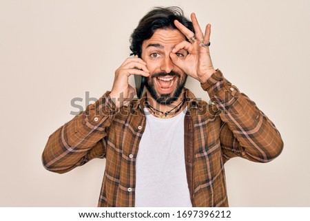 Young handsome hispanic man having a conversation talking on smartphone with happy face smiling doing ok sign with hand on eye looking through fingers