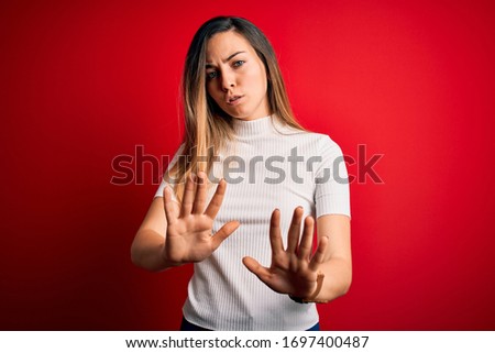 Beautiful blonde woman with blue eyes wearing casual white t-shirt over red background Moving away hands palms showing refusal and denial with afraid and disgusting expression. Stop and forbidden.