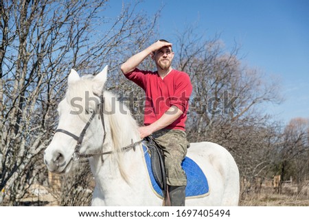 A man with a beard in a red sweater sits on a white horse against a blue sky and looks into the distance, covering his eyes with his hand from the sun.