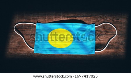 Palau National Flag at medical, surgical, protection mask on black wooden background. Coronavirus Covid–19, Prevent infection, illness or flu. State of Emergency, Lockdown...