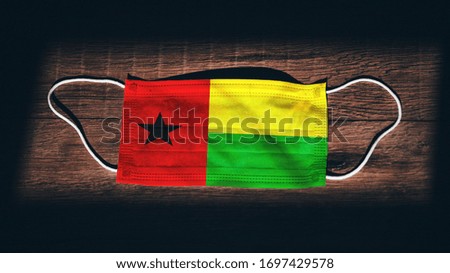 Guinea-Bissau National Flag at medical, surgical, protection mask on black wooden background. Coronavirus Covid–19, Prevent infection, illness or flu. State of Emergency, Lockdown...