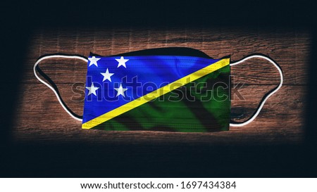Solomon Islands National Flag at medical, surgical, protection mask on black wooden background. Coronavirus Covid–19, Prevent infection, illness or flu. State of Emergency, Lockdown