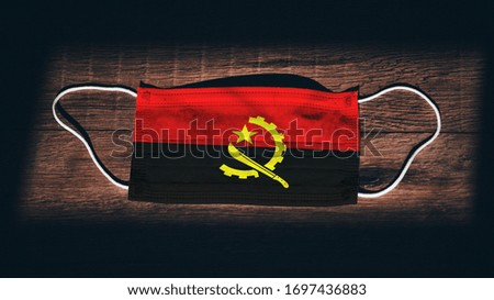 Angola National Flag at medical, surgical, protection mask on black wooden background. Coronavirus Covid–19, Prevent infection, illness or flu concept photo. State of Emergency, Lockdown