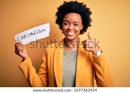 African American afro businesswoman with curly hair holding paper with capitalism message happy with big smile doing ok sign, thumb up with fingers, excellent sign