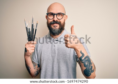 Handsome bald artist man with beard and tattoo painting using painter brushes happy with big smile doing ok sign, thumb up with fingers, excellent sign