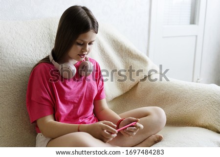 Cheerful girl in pink headphones and a red T-shirt sits with a phone in her hand. The concept of gadget and children's shopping online, home distance learning