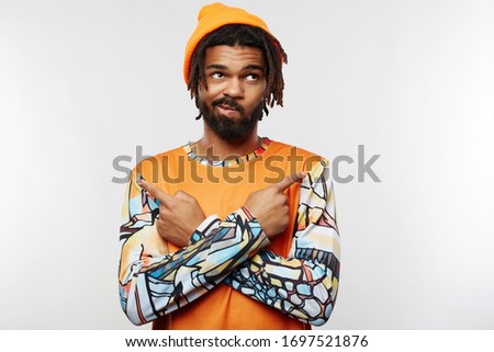 Displeased young handsome dark skinned guy with beard twisting his mouth while looking upwards and pointing in different sides with raised forefingers, isolated over white background